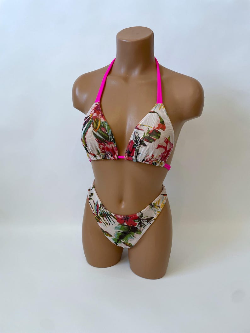 May include: A two-piece bikini set with a floral pattern. The bikini top has a halter neck with bright pink straps. The bottoms are high-waisted. The floral pattern includes red, green, and yellow flowers on a cream background.