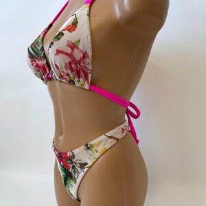 May include: A two-piece bikini set featuring a floral pattern with red, pink, and green accents on a white background. The bikini top has a triangle design with hot pink straps, and the bottoms are a matching high-cut style.