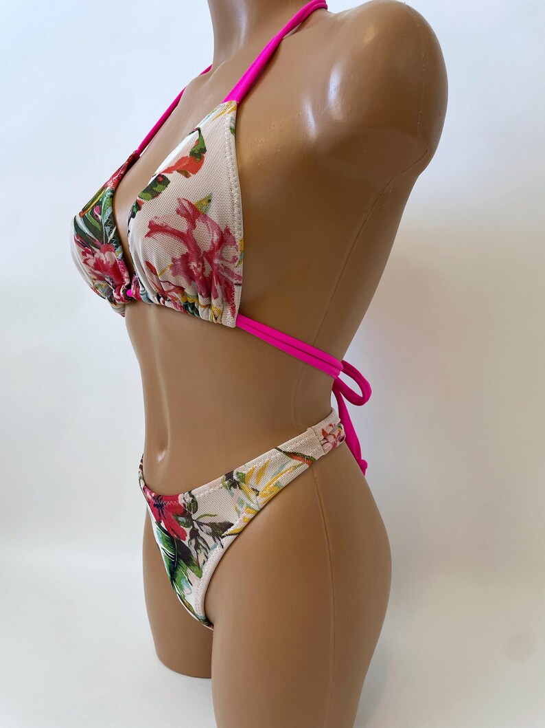 May include: A two-piece bikini set featuring a floral pattern with red, pink, and green accents on a white background. The bikini top has a triangle design with hot pink straps, and the bottoms are a matching high-cut style.