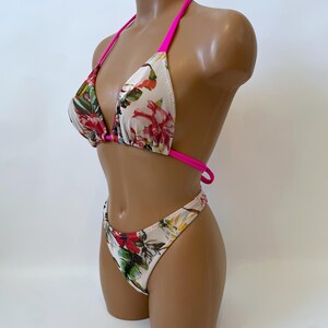 May include: A two-piece bikini set with a floral pattern. The bikini top has a triangle design with hot pink straps. The bottoms are high-cut with a matching floral print. The floral pattern includes red, green, and yellow flowers on a white background.