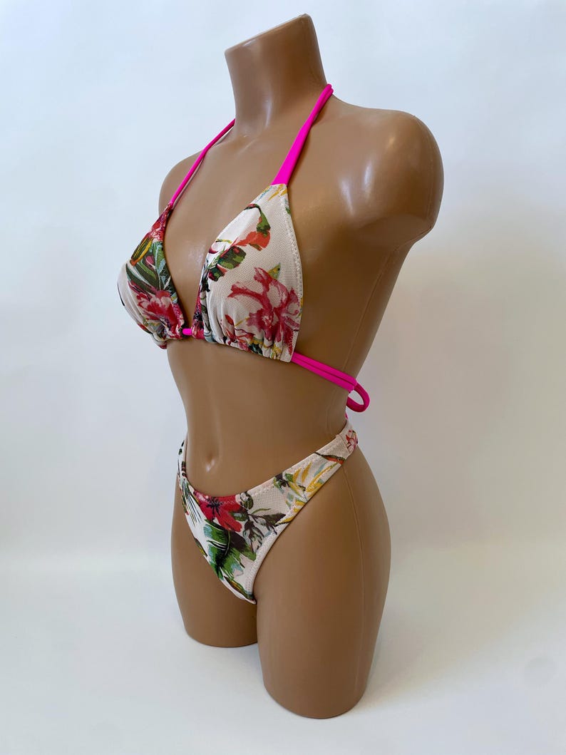 May include: A two-piece bikini set with a floral pattern. The bikini top has a triangle design with hot pink straps. The bottoms are high-cut with a matching floral print. The floral pattern includes red, green, and yellow flowers on a white background.