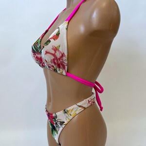May include: A two-piece bikini set with a floral pattern. The bikini top is a triangle style with hot pink straps. The bottoms are a matching high-cut style. The floral pattern includes red, pink, and green flowers on a cream background.