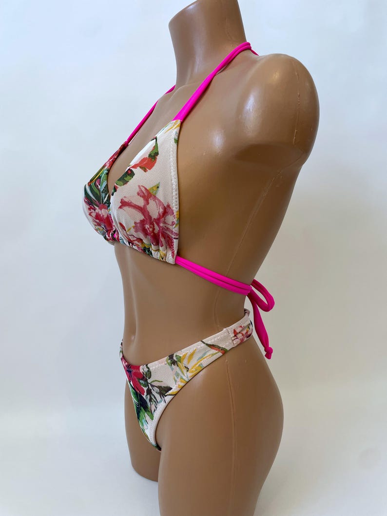 May include: A two-piece bikini set with a floral pattern. The bikini top is a triangle style with hot pink straps. The bottoms are a matching high-cut style. The floral pattern includes red, pink, and green flowers on a cream background.