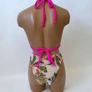 May include: A two-piece swimsuit displayed on a mannequin. The top is a halter style with bright pink ties. The bottoms are high-waisted with a floral pattern of green, yellow, and orange leaves on a cream background. The ties are bright pink.