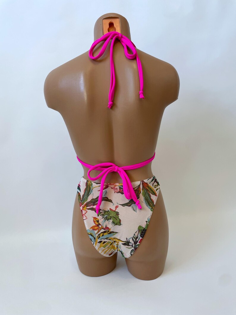 May include: A two-piece swimsuit displayed on a mannequin. The top is a halter style with bright pink ties. The bottoms are high-waisted with a floral pattern of green, yellow, and orange leaves on a cream background. The ties are bright pink.