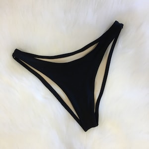 May include: Black bikini bottoms with a high-cut leg design. The bottoms have a solid black exterior and a light beige interior lining. The garment is displayed on a white, fluffy surface.
