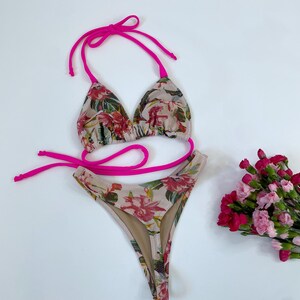 May include: A two-piece bikini set with a floral pattern in shades of pink, red, and green on a beige background. The bikini top and bottom are trimmed with bright pink ties. A small bouquet of pink and red flowers is to the side.