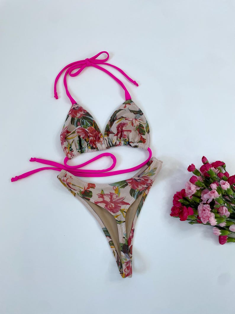 May include: A two-piece bikini set with a floral pattern in shades of pink, red, and green on a beige background. The bikini top and bottom are trimmed with bright pink ties. A small bouquet of pink and red flowers is to the side.