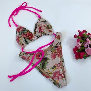 May include: A two-piece bikini set with a floral pattern in shades of pink, red, and green on a beige background. The bikini top and bottom are trimmed with bright pink ties. A small bouquet of pink flowers is visible.