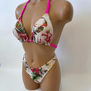 May include: A two-piece bikini set with a floral pattern. The bikini top has a triangle design with hot pink straps. The bottoms are high-cut with a matching floral print. The floral pattern includes red, green, and white flowers and leaves.