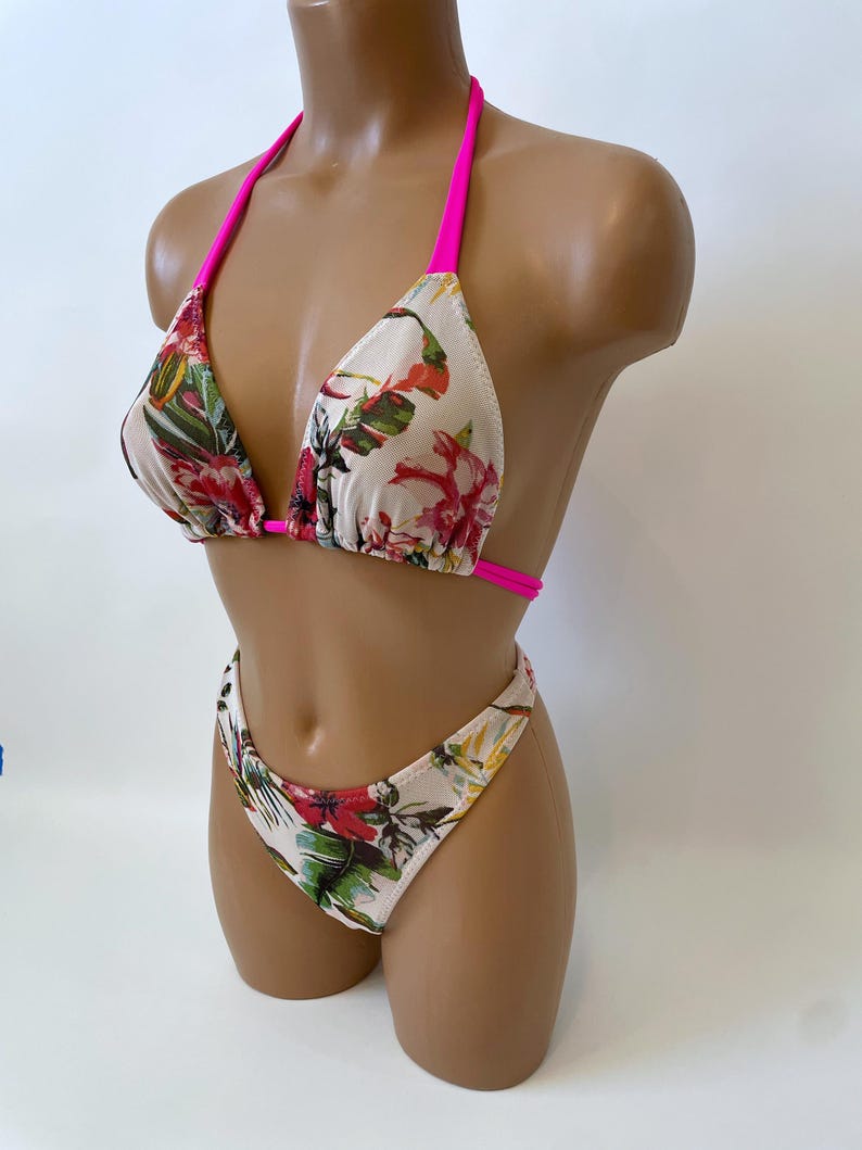 May include: A two-piece bikini set with a floral pattern. The bikini top has a triangle design with hot pink straps. The bottoms are high-cut with a matching floral print. The floral pattern includes red, green, and white flowers and leaves.