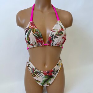 May include: A two-piece bikini set with a floral pattern. The bikini top has a halter neck design with bright pink straps. The bottoms are high-cut. The fabric has a cream background with red, green, and yellow floral print.