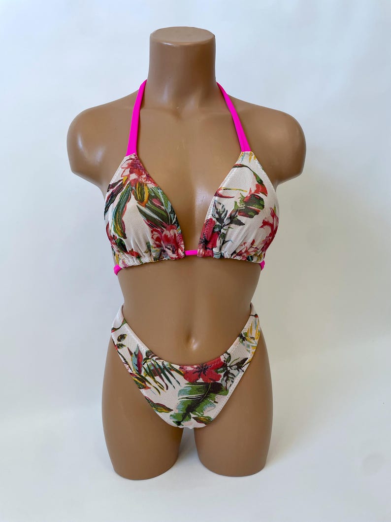 May include: A two-piece bikini set with a floral pattern. The bikini top has a halter neck design with bright pink straps. The bottoms are high-cut. The fabric has a cream background with red, green, and yellow floral print.