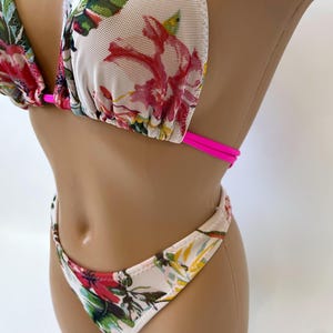 May include: A floral print bikini set featuring a triangle top and a low-rise bottom. The bikini has a white mesh base with a vibrant pattern of red, pink, and green flowers. The straps are a bright pink.