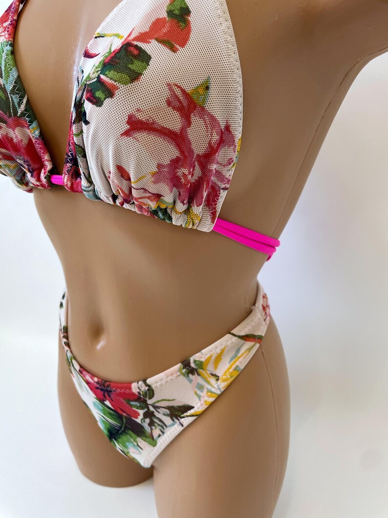 May include: A floral print bikini set featuring a triangle top and a low-rise bottom. The bikini has a white mesh base with a vibrant pattern of red, pink, and green flowers. The straps are a bright pink.