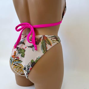 May include: A two-piece swimsuit with a floral pattern. The bikini top and bottoms feature a cream-colored background with colorful botanical designs. The straps and ties are a bright pink color.