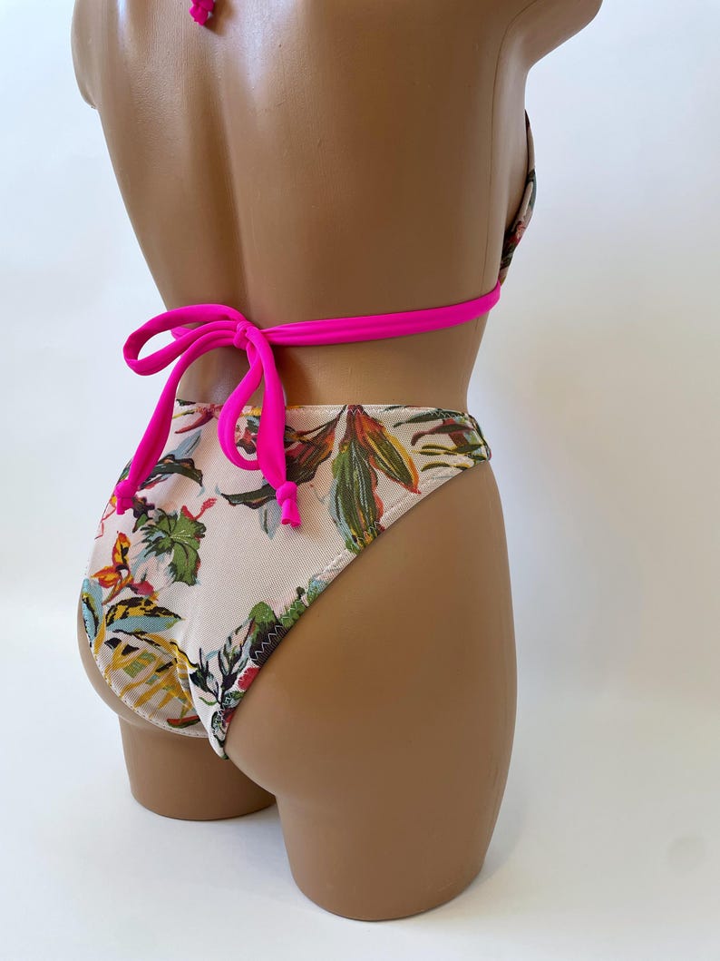 May include: A two-piece swimsuit with a floral pattern. The bikini top and bottoms feature a cream-colored background with colorful botanical designs. The straps and ties are a bright pink color.