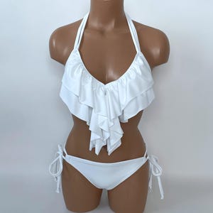 May include: A white bikini set displayed on a mannequin. The top is a halter style with layered ruffles. The bottoms are a classic tie-side design. The swimwear is made of a smooth, solid white fabric, perfect for a beach or pool day.