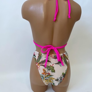 May include: A two-piece swimsuit set on a mannequin. The top is a bright pink halter style, and the bottoms are high-waisted with a floral pattern in shades of green, yellow, and brown on a cream background. The swimsuit is tied at the neck and back.