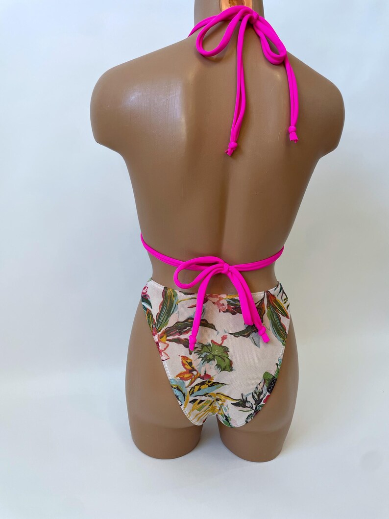 May include: A two-piece swimsuit set on a mannequin. The top is a bright pink halter style, and the bottoms are high-waisted with a floral pattern in shades of green, yellow, and brown on a cream background. The swimsuit is tied at the neck and back.