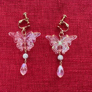 May include: A pair of clip-on earrings featuring a butterfly design. The earrings have a gold-colored clip and a clear, iridescent butterfly shape. Below the butterfly is a small, sparkly ball and a teardrop-shaped crystal. The background is a textured red fabric.