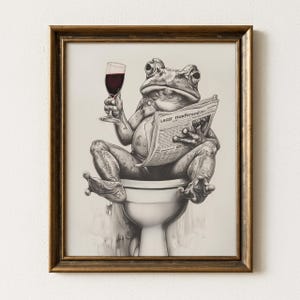 May include: A framed black and white print depicts a frog sitting on a toilet, holding a glass of red wine and reading a newspaper. The newspaper has text. The frame is a warm brown color.