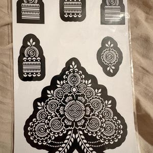May include: A sheet of black adhesive stencils featuring intricate floral and geometric patterns. The design includes four smaller arched motifs with horizontal lines and swirls, one smaller circular motif, and one large central floral arrangement.