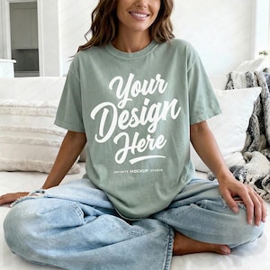 May include: Sage green t-shirt with the text "Your Design Here" in a white script font. The shirt is part of a clothing mockup, and the text "INFINITE MOCKUP STUDIO" is printed below the main text. The model is wearing light blue jeans.