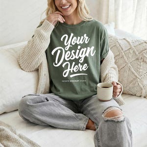 May include: An olive green t-shirt with the text "Your Design Here" in white script. The shirt is worn with a beige cardigan and gray jeans. A beige mug is held in the hand. The shirt is from Infinite Mockup Studio.