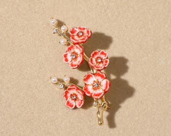 Puede incluir: Un broche de metal dorado con forma de rama floreciente. El diseño presenta cinco flores de esmalte rojo y blanco con pequeños cristales transparentes en el centro, acentuadas por diminutas perlas blancas a lo largo del tallo y las puntas.