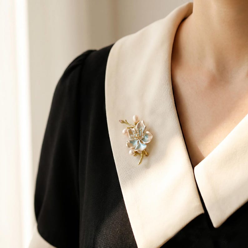 Puede incluir: Un broche de metal dorado con forma de flor y p&eacute;talos de esmalte azul claro y blanco. El dise&ntilde;o floral presenta peque&ntilde;os detalles en forma de perla blanca en los tallos y el centro, prendido en una prenda negra con un cuello ancho de color crema.