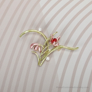 Puede incluir: Un broche floral con tallos y hojas de color verde claro, dos capullos de flores rosas con centros rojos y dos peque&ntilde;os acentos esf&eacute;ricos blancos. El dise&ntilde;o es delicado y bot&aacute;nico, con un acabado pulido. El texto AlyssaCustomization.etsy.com es visible.