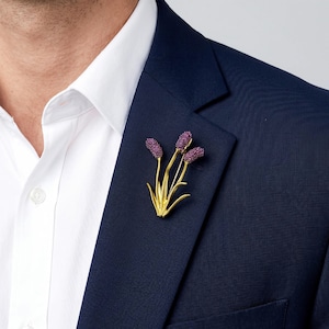 Puede incluir: Un broche de metal dorado con forma de racimo de tres tallos florales. Cada tallo presenta una cabeza redondeada y texturizada de color morado intenso en la parte superior, sostenida por tallos dorados finos y lisos con hojas puntiagudas.