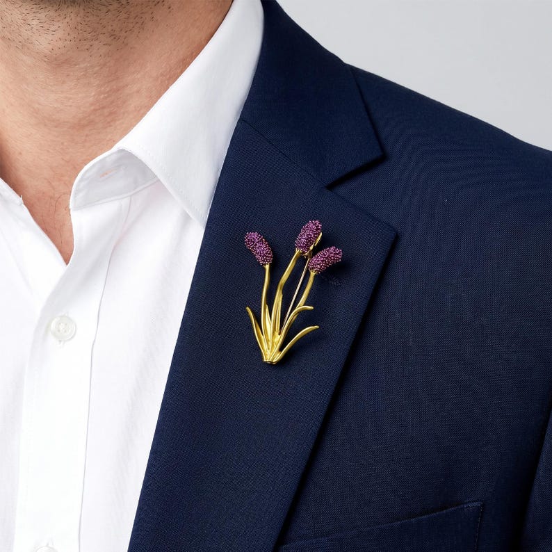 Puede incluir: Un broche de metal dorado con forma de racimo de tres tallos florales. Cada tallo presenta una cabeza redondeada y texturizada de color morado intenso en la parte superior, sostenida por tallos dorados finos y lisos con hojas puntiagudas.