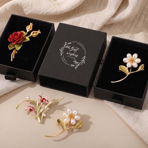 Puede incluir: Cuatro broches florales con tallos y hojas doradas. Un broche tiene una flor roja, otro una flor de perlas blancas y dos tienen detalles florales rosas y blancos. Dos broches est&aacute;n en cajas negras con el texto Best wishes 17/9/2026.