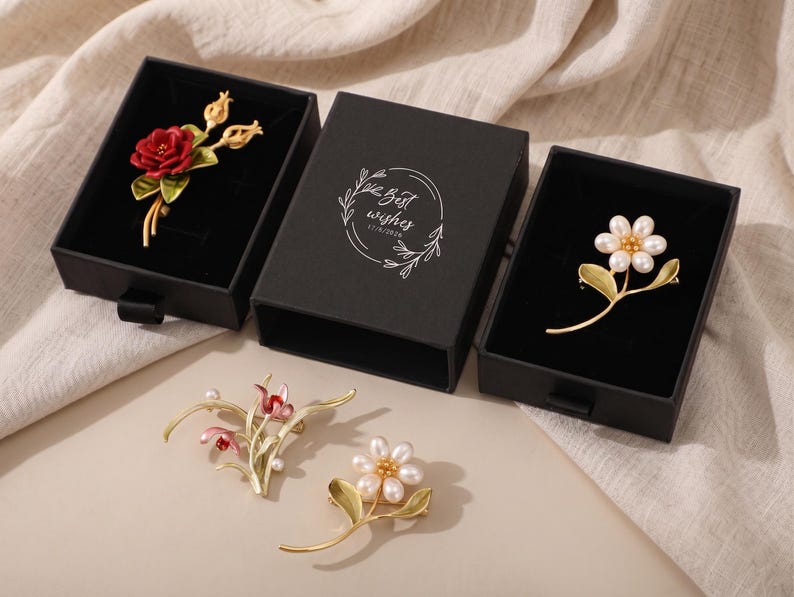 Puede incluir: Cuatro broches florales con tallos y hojas doradas. Un broche tiene una flor roja, otro una flor de perlas blancas y dos tienen detalles florales rosas y blancos. Dos broches est&aacute;n en cajas negras con el texto Best wishes 17/9/2026.
