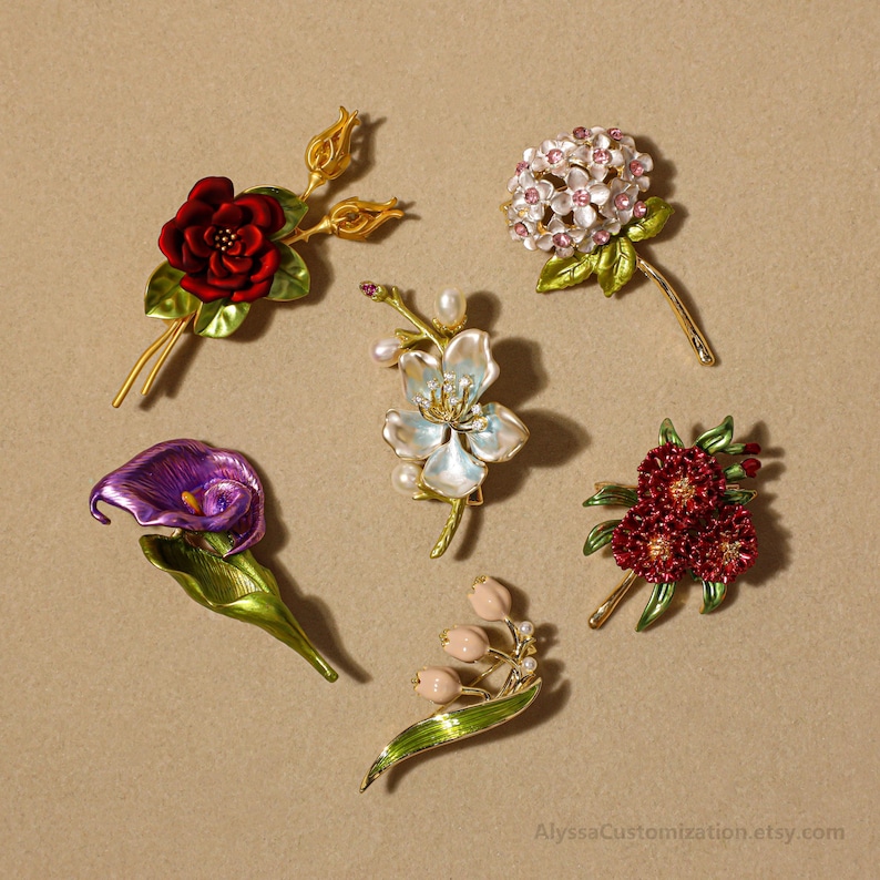 Puede incluir: Una colecci&oacute;n de seis broches florales que incluye una rosa roja, una hortensia blanca con cristales rosas, un lirio de agua morado, una flor blanca iridiscente con perlas, una flor roja en racimo y lirios del valle rosa p&aacute;lido con hojas verdes.