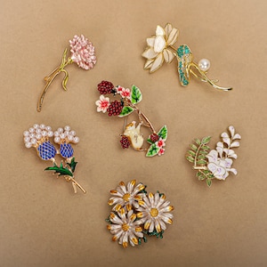 Puede incluir: Seis broches de flores de esmalte con detalles dorados. Los broches presentan una variedad de flores, incluyendo un clavel rosa, un loto blanco, una rama de bayas rojas, un cardo azul, una glicinia blanca y un grupo de margaritas blancas.