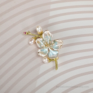 Puede incluir: Un broche dorado con forma de rama floreciente con p&eacute;talos de esmalte azul claro y blanco, tres perlas blancas, peque&ntilde;as piedras transparentes en el centro y una diminuta piedra rosa en la punta. El texto AlyssaCustomization.etsy.com es visible.