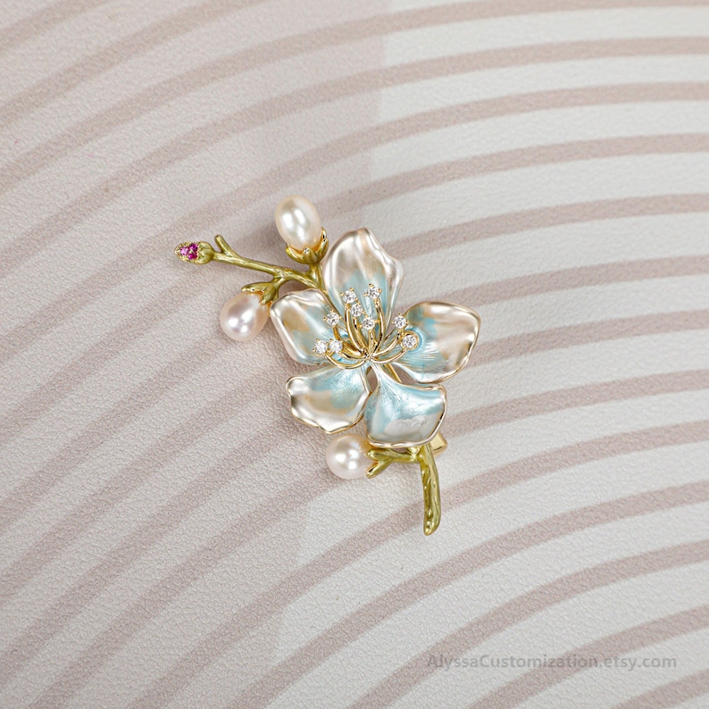 Puede incluir: Un broche dorado con forma de rama floreciente con p&eacute;talos de esmalte azul claro y blanco, tres perlas blancas, peque&ntilde;as piedras transparentes en el centro y una diminuta piedra rosa en la punta. El texto AlyssaCustomization.etsy.com es visible.