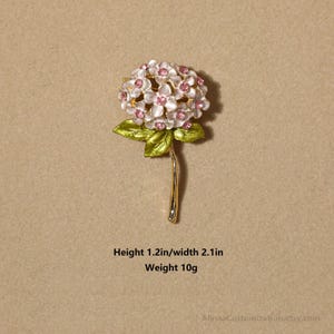 Puede incluir: Un broche de metal dorado con forma de flor de hortensia con p&eacute;talos blancos, centros de pedrer&iacute;a rosa y hojas verdes. El broche mide 3,05 cm de alto, 5,33 cm de ancho y pesa 10 gramos, tal como se indica en el texto impreso.