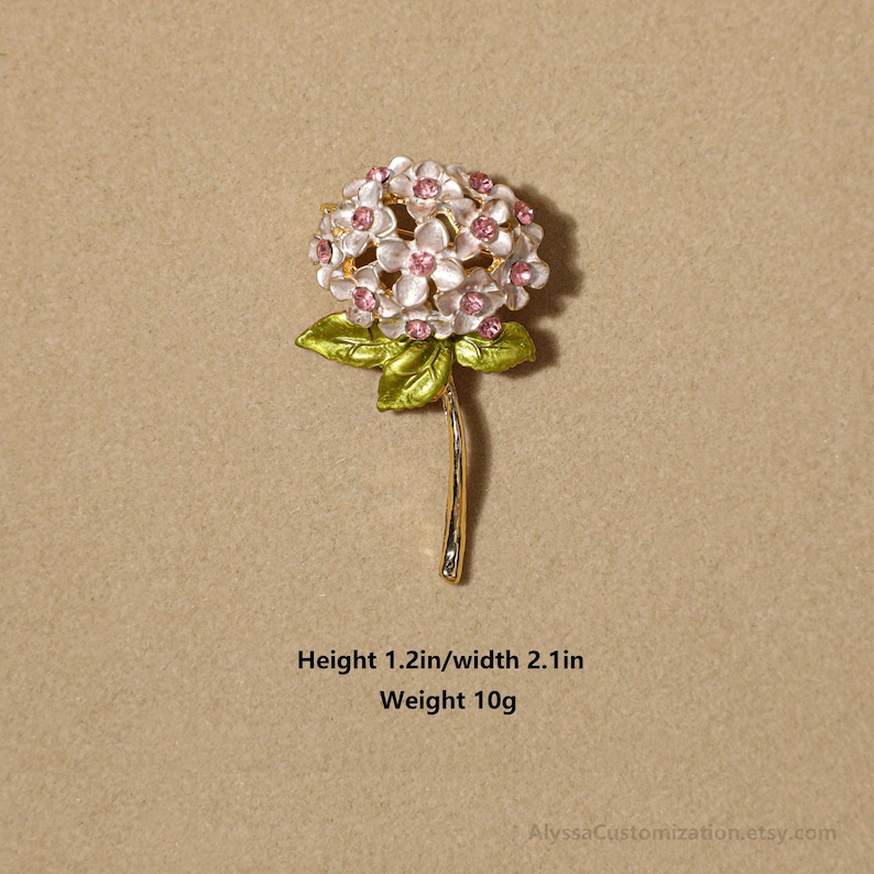 Puede incluir: Un broche de metal dorado con forma de flor de hortensia con p&eacute;talos blancos, centros de pedrer&iacute;a rosa y hojas verdes. El broche mide 3,05 cm de alto, 5,33 cm de ancho y pesa 10 gramos, tal como se indica en el texto impreso.