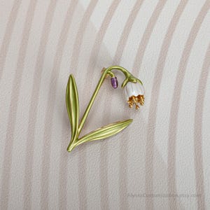 Puede incluir: Un broche de metal con forma de flor de campanilla con tallo y hojas verdes, una flor blanca en forma de campana, estambres dorados y un peque&ntilde;o capullo morado. El texto AlyssaCustomization.etsy.com es visible en la esquina inferior derecha.