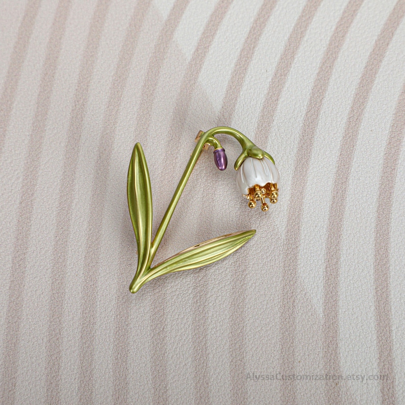 Puede incluir: Un broche de metal con forma de flor de campanilla con tallo y hojas verdes, una flor blanca en forma de campana, estambres dorados y un peque&ntilde;o capullo morado. El texto AlyssaCustomization.etsy.com es visible en la esquina inferior derecha.