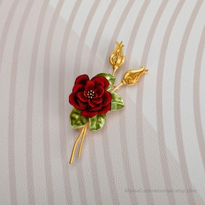 Puede incluir: Un broche de metal en tono dorado que presenta una flor de rosa esmaltada en rojo intenso con un centro amarillo, acompa&ntilde;ada de dos capullos de rosa dorados y hojas esmaltadas en verde. Los tallos son delgados y dorados, extendi&eacute;ndose.
