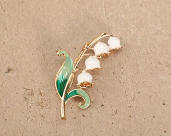Op de afbeelding: Een broche van goudkleurig metaal in de vorm van een lelietje-van-dalen. Het ontwerp bevat drie kleine, ronde, wit geëmailleerde klokvormige bloemetjes aan een gebogen stengel, vergezeld door twee langwerpige groene bladeren met kleurverloop.