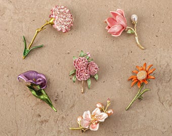 Puede incluir: Una colección de seis broches florales con diversos diseños, incluyendo un clavel rosa, una magnolia rosa con una perla, un lirio de agua morado, un racimo de flores rosas, una margarita naranja y una rama de flores blancas con perlas.