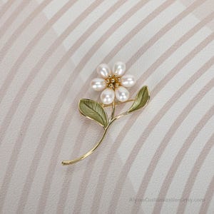 Puede incluir: Un broche de metal dorado con forma de flor con cinco perlas blancas ovaladas como p&eacute;talos. El centro de la flor presenta peque&ntilde;as cuentas doradas. El tallo incluye dos hojas esmaltadas en verde con detalles de nervaduras visibles.