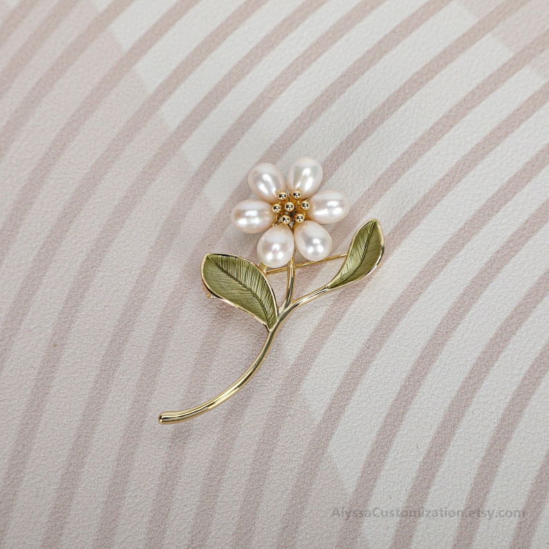 Puede incluir: Un broche de metal dorado con forma de flor con cinco perlas blancas ovaladas como p&eacute;talos. El centro de la flor presenta peque&ntilde;as cuentas doradas. El tallo incluye dos hojas esmaltadas en verde con detalles de nervaduras visibles.