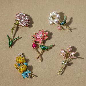 Könnte beinhalten: Eine Auswahl an floralen Broschen in verschiedenen Designs. Die Broschen weisen detaillierte Emaillearbeiten in Farben wie Rosa, Weiß, Gelb und Grün mit Perlenakzenten auf. Einige Designs beinhalten kleine Vogelfiguren.